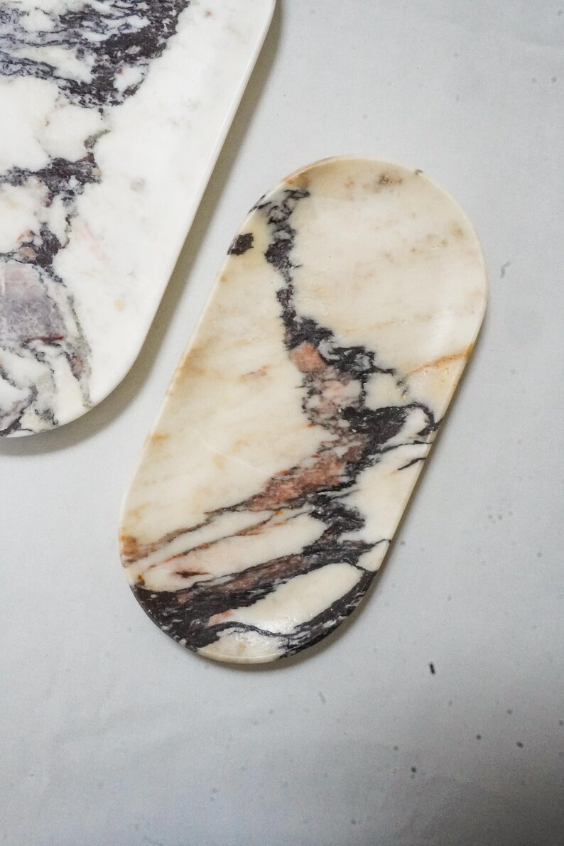 May include: Two marble trays with a natural stone pattern. The trays have a cream-colored base with dark brown and reddish-brown veining. One tray is oval-shaped, and the other is a long, rectangular shape.