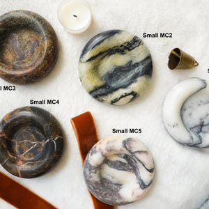 May include: Five circular stone dishes with varied marble patterns labeled Small MC1 through Small MC5. The dishes feature colors including dark brown, black, white, and yellow-toned veining. A small brass bell and a candle are also visible.