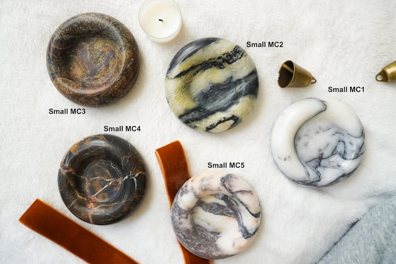 May include: Five circular stone dishes with varied marble patterns labeled Small MC1 through Small MC5. The dishes feature colors including dark brown, black, white, and yellow-toned veining. A small brass bell and a candle are also visible.