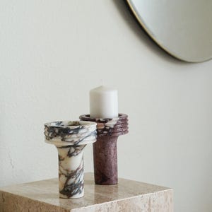 Puede incluir: Dos portavelas cil&iacute;ndricos de piedra con partes superiores acanaladas. Un soporte presenta una base blanca con vetas gris oscuro y marrones, mientras que el otro es de piedra color granate oscuro s&oacute;lido. Una vela blanca descansa en el soporte granate.