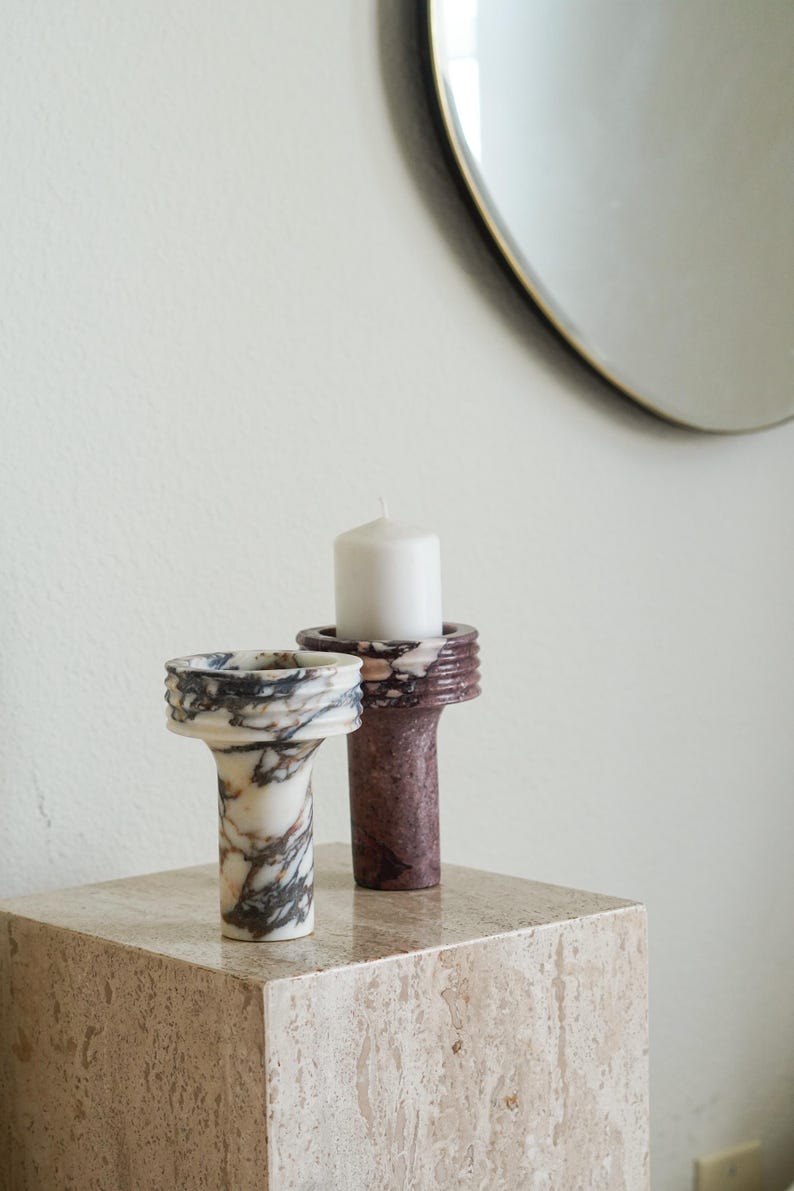 Puede incluir: Dos portavelas cil&iacute;ndricos de piedra con partes superiores acanaladas. Un soporte presenta una base blanca con vetas gris oscuro y marrones, mientras que el otro es de piedra color granate oscuro s&oacute;lido. Una vela blanca descansa en el soporte granate.