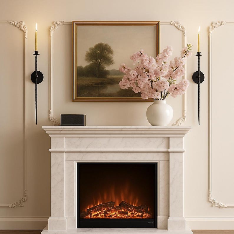 May include: A white marble fireplace with a black electric firebox. Above the fireplace is a framed landscape painting, a white vase with pink flowers, and a black box. Two black sconces with lit candles flank the painting.