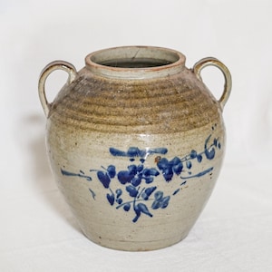 May include: A stoneware jar with two side handles, featuring a light beige body with a darker brown glazed rim and a hand-painted blue floral design on the front. The vessel has a wide circular opening and a rounded, bulbous shape.