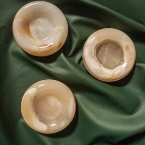 May include: Three circular, polished stone bowls with a smooth, glossy finish. Each bowl features a translucent, pale beige and cream color palette with subtle, swirling brown and white mineral inclusions throughout the natural stone material.