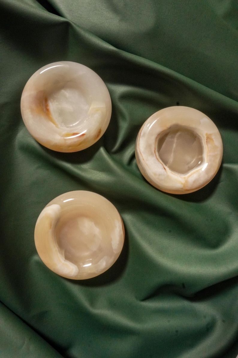May include: Three circular, polished stone bowls with a smooth, glossy finish. Each bowl features a translucent, pale beige and cream color palette with subtle, swirling brown and white mineral inclusions throughout the natural stone material.