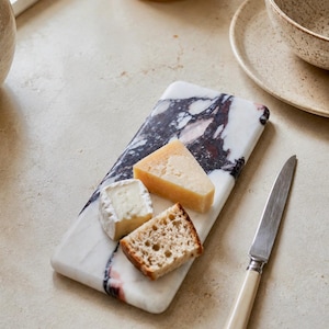 May include: A rectangular white marble serving board with prominent dark purple and black veining. The board holds a wedge of yellow cheese, a small piece of white soft cheese, and a slice of crusty bread, accompanied by a silver knife with a cream handle.