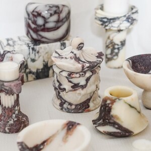 Puede incluir: Una colecci&oacute;n de objetos decorativos hechos de m&aacute;rmol blanco con vetas de color p&uacute;rpura oscuro y negro, que incluye un tarro con tapa, varios cuencos peque&ntilde;os, portavelas estilo pilar y un bloque rectangular, todos con superficies pulidas.