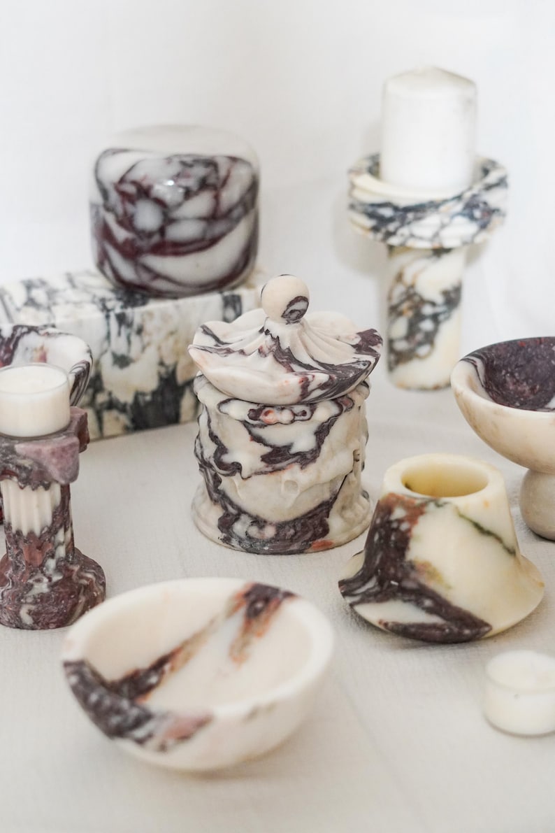 Puede incluir: Una colecci&oacute;n de objetos decorativos hechos de m&aacute;rmol blanco con vetas de color p&uacute;rpura oscuro y negro, que incluye un tarro con tapa, varios cuencos peque&ntilde;os, portavelas estilo pilar y un bloque rectangular, todos con superficies pulidas.