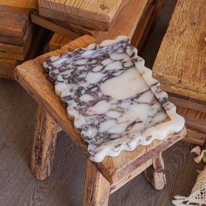 May include: A rectangular marble tray featuring a scalloped edge and a polished surface with a white and deep purple veined pattern. The tray is positioned on a small, rustic wooden stool with four thick, textured legs and a flat, weathered top.