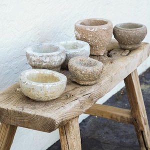 May include: A collection of five small, weathered stone mortars with rough, textured surfaces in shades of gray, beige, and tan. These heavy, circular vessels feature thick walls and open centers, arranged in a cluster on a rustic wooden bench.