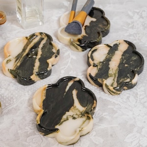 May include: Four clover-shaped trays made of polished stone with a marbled pattern of dark green, black, and cream. One tray holds two makeup brushes with grey bristles and gold-toned ferrules. A clear glass bottle with a silver cap is visible.