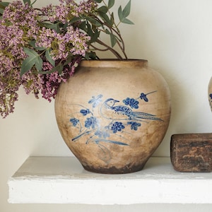 May include: A round, tan-colored ceramic jar with a weathered, mottled texture. The jar features a blue painted illustration of a bird perched on a flowering branch. A cluster of purple flowers with green leaves is placed inside the jar opening.