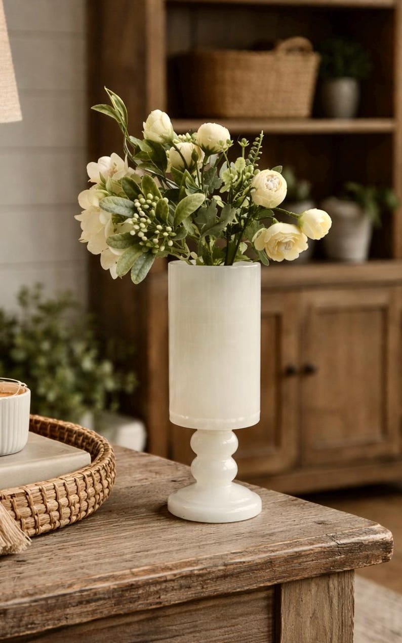 Puede incluir: Un jarr&oacute;n cil&iacute;ndrico blanco con base de pedestal, que contiene un ramo de flores y vegetaci&oacute;n color crema. El jarr&oacute;n est&aacute; hecho de un material liso y opaco y se encuentra sobre una superficie de madera. La est&eacute;tica general es una mezcla de r&uacute;stico y elegante.