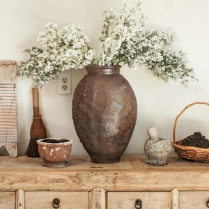May include: A rustic, brown, textured vase filled with white flowers sits on a wooden cabinet. Other items on the cabinet include a small bowl, a mortar and pestle, a small broom, and a wicker basket.