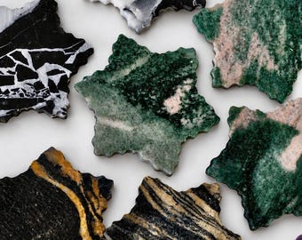 Puede incluir: Una colección de posavasos de piedra con forma de estrella que presentan varios patrones y colores naturales, incluyendo mármol blanco y negro, verde intenso con inclusiones rosadas y negro con vetas doradas.