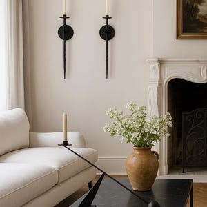 May include: A living room scene featuring a black marble coffee table, a white sofa, and two black wall sconces with candles. A vase with white flowers sits on the table. A painting and fireplace are in the background.