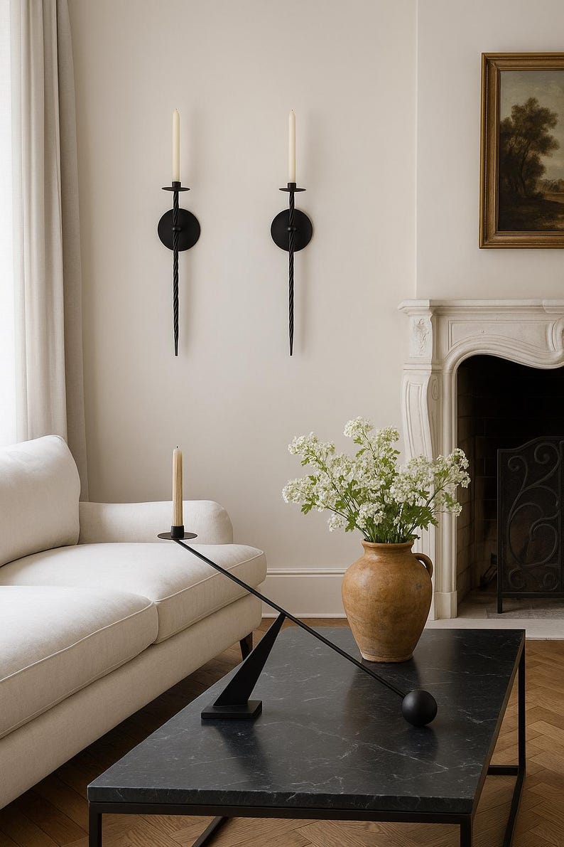 May include: A living room scene featuring a black marble coffee table, a white sofa, and two black wall sconces with candles. A vase with white flowers sits on the table. A painting and fireplace are in the background.