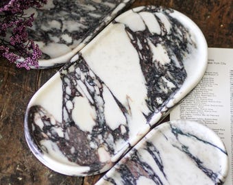 May include: Three oval-shaped trays crafted from white marble with prominent dark purple and black veining. The trays feature smooth, rounded edges and a polished surface, showcasing the natural stone patterns in varying sizes.
