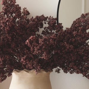 May include: A beige ceramic vase with a ruffled rim holds a large arrangement of dried burgundy flowers. The flowers have small, clustered buds. A black, round mirror is partially visible in the background.