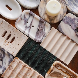 May include: A collection of stone soap dishes and trays in various shapes, colors, and textures, including white, grey, beige, green, and purple marble, travertine, and onyx, with some featuring drainage holes and others having a ribbed surface.