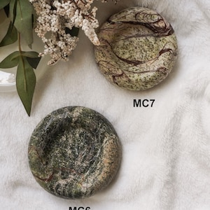 May include: Two round stone dishes with polished surfaces. The top dish, labeled MC7, features a light green base with dark brown swirling veins. The bottom dish, labeled MC6, displays a deep forest green color with intricate white and dark veining.