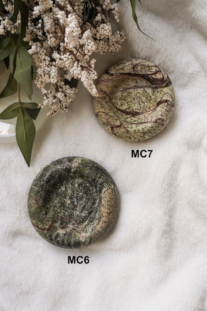 May include: Two round stone dishes with polished surfaces. The top dish, labeled MC7, features a light green base with dark brown swirling veins. The bottom dish, labeled MC6, displays a deep forest green color with intricate white and dark veining.