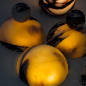 Puede incluir: Una colecci&oacute;n de objetos de piedra transl&uacute;cidos en forma de c&uacute;pula con vetas de color marr&oacute;n oscuro y negro. Cada pieza emite un c&aacute;lido resplandor dorado interno, resaltando los patrones naturales de la piedra. Algunas c&uacute;pulas tienen un remate esf&eacute;rico.