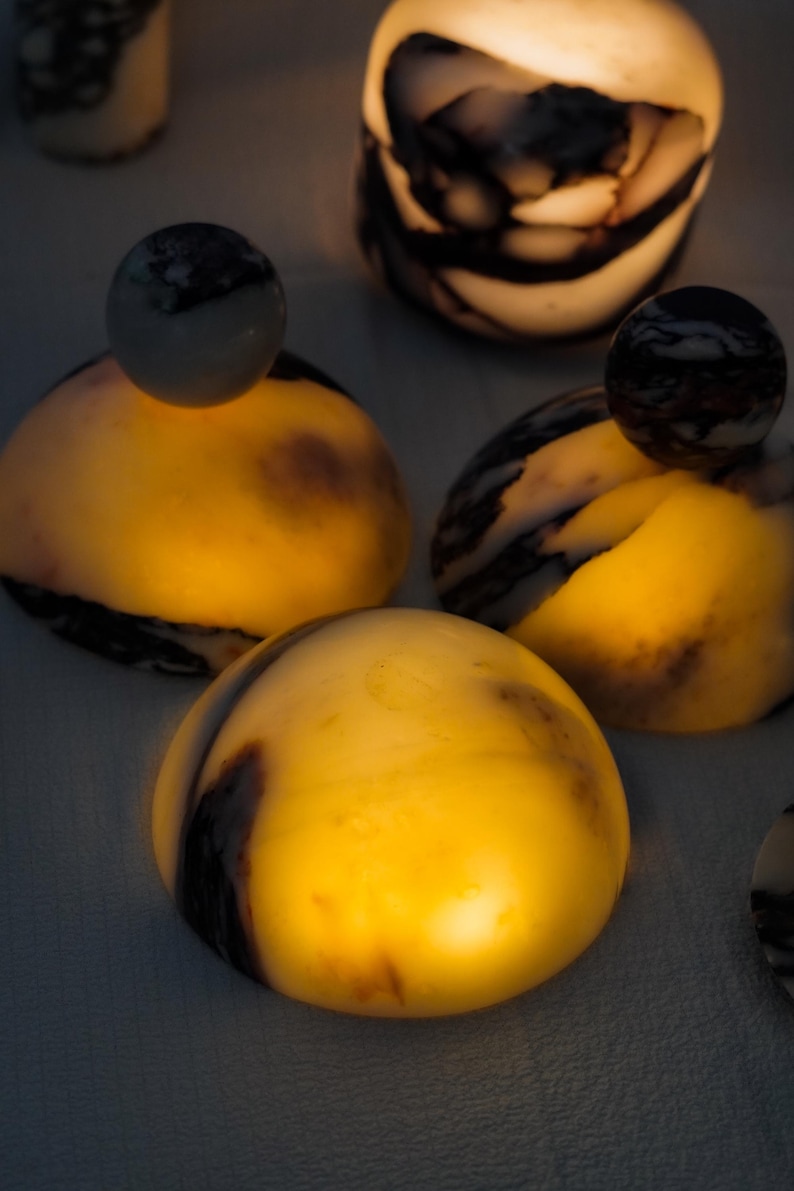 Puede incluir: Una colecci&oacute;n de objetos de piedra transl&uacute;cidos en forma de c&uacute;pula con vetas de color marr&oacute;n oscuro y negro. Cada pieza emite un c&aacute;lido resplandor dorado interno, resaltando los patrones naturales de la piedra. Algunas c&uacute;pulas tienen un remate esf&eacute;rico.