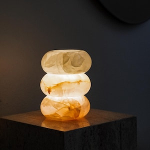 May include: A table lamp composed of three stacked, rounded, translucent stone rings. The lamp emits a warm, soft glow from within, highlighting the natural veining and creamy, amber-toned textures of the polished stone material throughout.