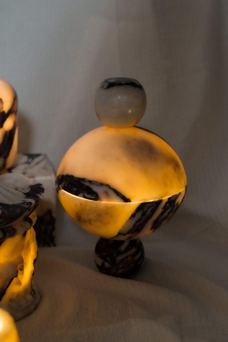 Puede incluir: Un recipiente esf&eacute;rico de piedra con un patr&oacute;n veteado transl&uacute;cido en color crema y marr&oacute;n oscuro, que presenta una base circular y una tapa redonda a juego con un peque&ntilde;o pomo, iluminado desde el interior para crear un efecto c&aacute;lido.