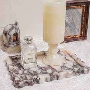 Puede incluir: Una bandeja decorativa de m&aacute;rmol con borde ondulado sostiene una vela grande, un frasco de perfume y una peque&ntilde;a casa de m&aacute;rmol. Una imagen enmarcada est&aacute; en el fondo. La bandeja y la casa tienen un patr&oacute;n blanco y burdeos.
