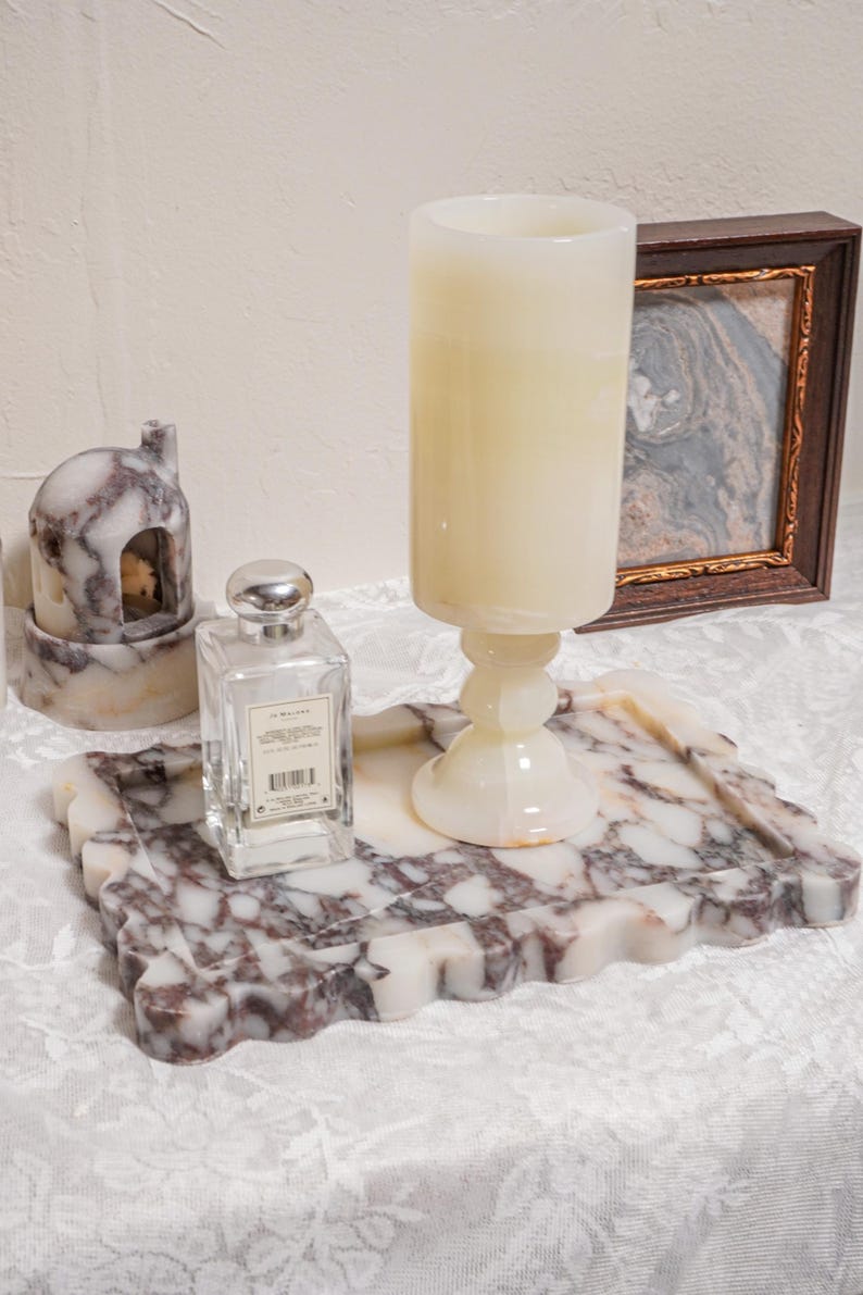 Puede incluir: Una bandeja decorativa de m&aacute;rmol con borde ondulado sostiene una vela grande, un frasco de perfume y una peque&ntilde;a casa de m&aacute;rmol. Una imagen enmarcada est&aacute; en el fondo. La bandeja y la casa tienen un patr&oacute;n blanco y burdeos.