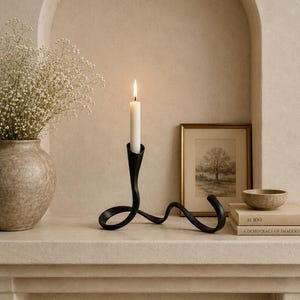 May include: A black metal candle holder with a lit white candle. The holder has a unique, flowing design. A vase with white flowers, a framed picture, a small bowl, and books are also on the shelf.