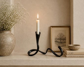 May include: A black metal candle holder with a lit white candle. The holder has a unique, flowing design. A vase with white flowers, a framed picture, a small bowl, and books are also on the shelf.