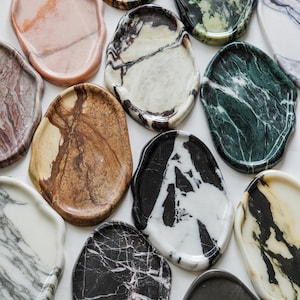May include: A collection of irregular, organic-shaped stone trays featuring various natural patterns and colors, including black, white, green, brown, and pink marble-like veining, arranged in a cluster to display their unique textures and finishes.
