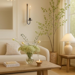 May include: A cozy living room scene with a light-colored sofa, wooden coffee table, and a vase of flowers. A lit candle and a book are on the table. A decorative light fixture hangs from the ceiling, and a tree is in the background.