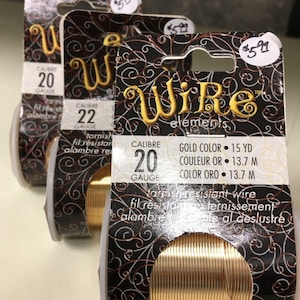 May include: Three spools of gold colored wire, labeled "Wire elements" with the text "Calibre 20 Gauge" and "Gold Color 15 yd, Couleur Or 13.7 m, Color Oro 13.7 m". The wire is described as "Varnish resistant wire, fil resistant, ternissement, alambre resistente al deslustre".