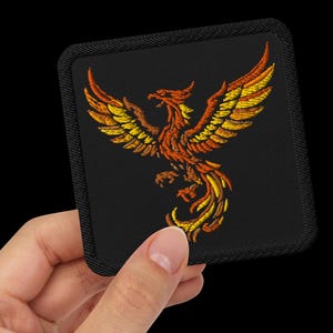 May include: A square-shaped embroidered patch featuring a phoenix with wings spread wide. The phoenix is stitched in vibrant shades of orange, red, and yellow thread, set against a solid black background with a black stitched border edge.