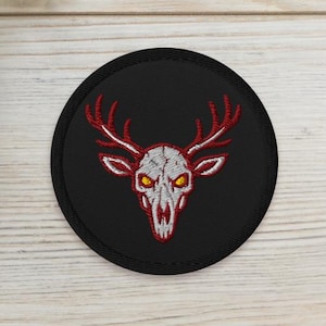 May include: A round black patch featuring a white deer skull with red antlers and yellow eyes. The skull is embroidered with red thread. Two small potted succulents are in the background.