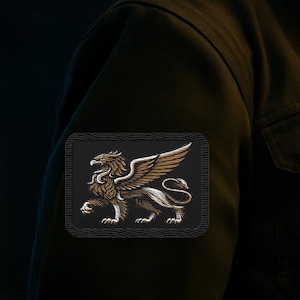May include: A rectangular patch featuring an embroidered gold and white griffin on a black background. The mythical creature is depicted in profile with wings spread, positioned on the sleeve of a dark olive green jacket with a visible chest pocket.