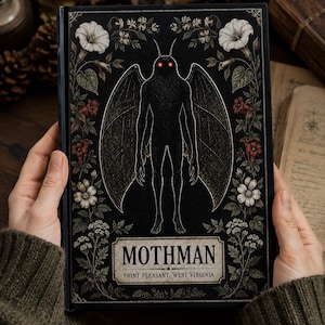 May include: A black hardcover book featuring a central illustration of the Mothman with glowing red eyes, surrounded by a detailed botanical border of white and red flowers. The text reads MOTHMAN POINT PLEASANT, WEST VIRGINIA.