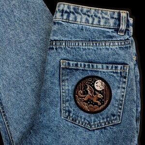 May include: A pair of blue denim jeans with a patch on the back pocket. The patch is a circular design with a brown bat flying in front of a full moon. The bat is outlined in white and has a brown body with white wings.