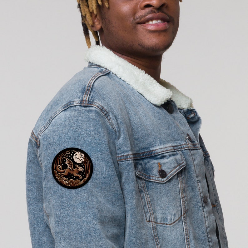 May include: A blue denim jacket with a brown and white embroidered patch of a dragon flying in front of a full moon. The patch is on the left sleeve of the jacket.