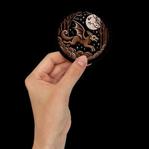 May include: A round sticker with a black background and a gold design. The design features a winged creature with a fox-like head flying through a forest scene with a full moon in the background.