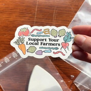 Może przedstawiać: Naklejka z napisem "Support Your Local Farmers" oraz ilustracjami warzyw i narzędzi ogrodniczych. Naklejka przedstawia pomidora, marchewkę, karczochy, szparagi, rzodkiewkę i robaka. Naklejka jest trzymana w dłoni.