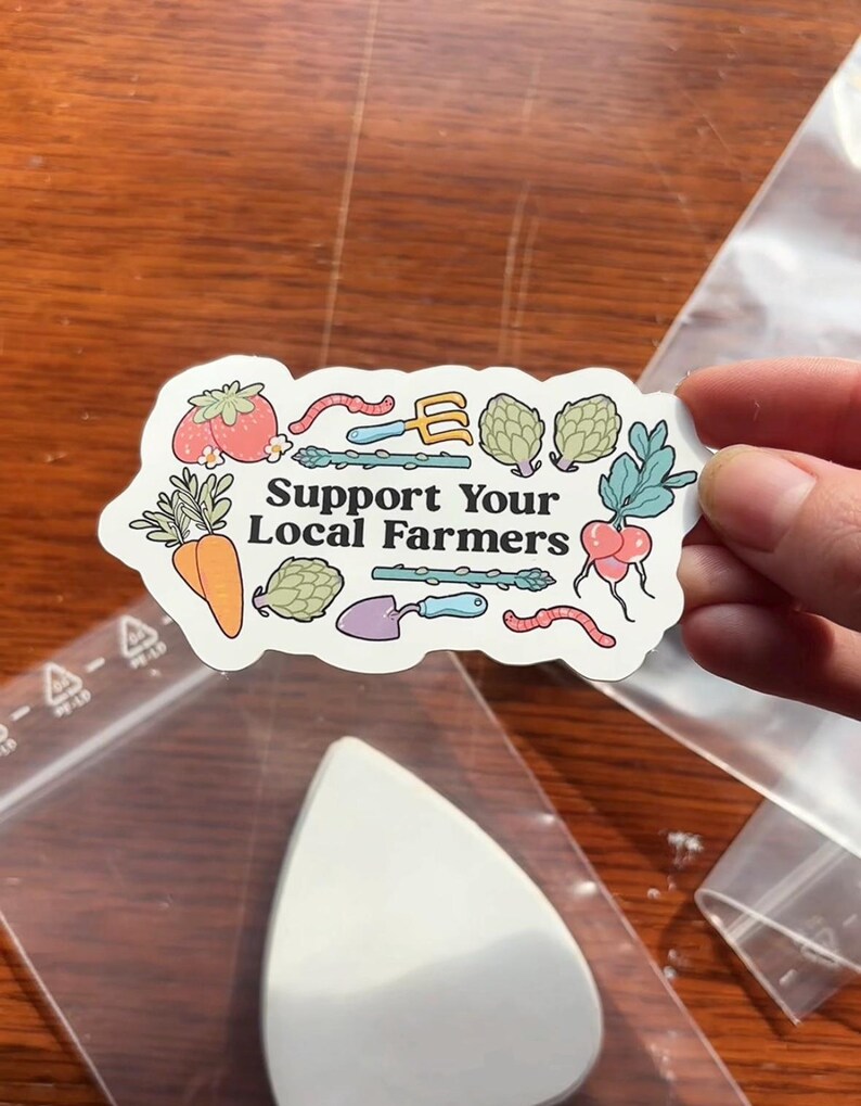 Może przedstawiać: Naklejka z napisem "Support Your Local Farmers" oraz ilustracjami warzyw i narzędzi ogrodniczych. Naklejka przedstawia pomidora, marchewkę, karczochy, szparagi, rzodkiewkę i robaka. Naklejka jest trzymana w dłoni.