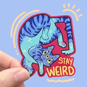 Może przedstawiać: Naklejka przedstawiająca niebieskiego kota z wieloma oczami w wykrzywionej pozie na czerwonym tle. Naklejka zawiera żółty napis "STAY WEIRD". Kot ma niebieskie paski i jest umieszczony na jasnoniebieskim tle.