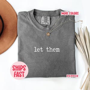 May include: A gray Comfort Colors t-shirt with the text "let them" in white. A gold necklace with a pendant rests on the shirt. The shirt is folded and displayed with a brown hat and dried plant stems.