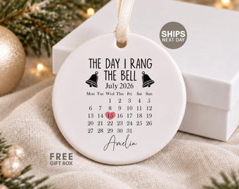 May include: A round white ceramic ornament with a ribbon hanger. The text reads THE DAY I RANG THE BELL, July 2026, and the name Amelia. It features a calendar for July 2026 with a pink heart highlighting the 15th and two black bell icons.