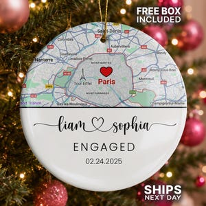 May include: A round white ceramic ornament featuring a map of Paris with a red heart icon over the city center. Below the map, the text reads Liam and Sophia, ENGAGED, and the date 02.24.2025 in black lettering. A gold string is attached to the top.